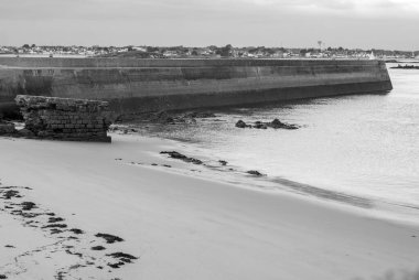 Büyük bir kaya duvarı ve küçük bir binası olan bir plajın siyah beyaz fotoğrafı. Gavres Yarımadası, Britanya, Fransa