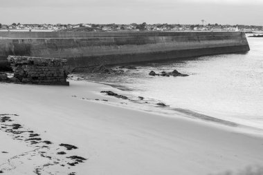 Arka planda büyük bir taş duvar olan bir plajın siyah beyaz fotoğrafı. Duvar taştan yapılmıştır ve suya yakındır. Kumsal boş ve su sakin. Fotoğrafta huzur var. Gavres Yarımadası, Britanya, Fransa