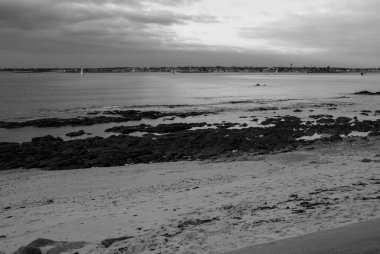 Kayalık bir kıyı şeridi olan bir plajın siyah beyaz fotoğrafı. Su sakin ve gökyüzü bulutlu. Gavres Yarımadası, Britanya, Fransa