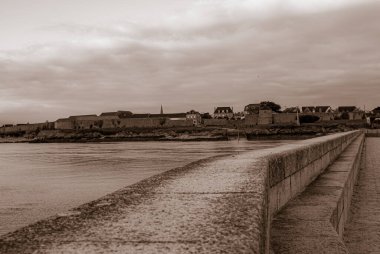 Taştan duvarı ve köprüsü olan bir sahil. Gökyüzü bulutlu ve su sakin. Gavres Yarımadası, Britanya, Fransa
