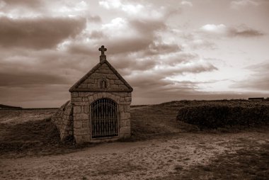 Üzerinde haç olan küçük bir taş bina. Bina bir tarla ile çevrili ve gökyüzü bulutlu. Gavres Yarımadası, Britanya, Fransa