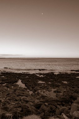 Büyük bir su kütlesi ve gökte bir ay olan bir sahil. Gökyüzü mavi ve turuncunun bir karışımıdır. Huzurlu ve huzurlu bir atmosfer yaratır. Gavres Yarımadası, Britanya, Fransa