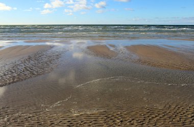 Ön planda ıslak kum ve sığ su, sakin bir okyanusa götürür. Çoğunlukla berrak bir gökyüzünün altında, dağınık bulutlarla, sakin bir kıyı sahnesini yakalar..
