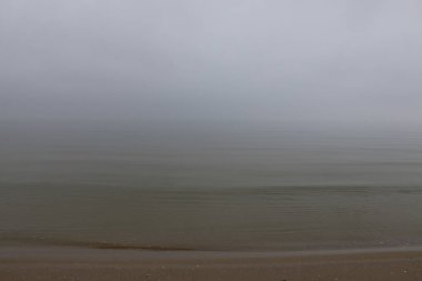 Sakin bulutlu deniz manzarası, kumlu bir sahilde hafif dalgalarla, sakin sular ufukta bulutlu bir gökyüzü ile birleşiyor, sükunet ve minimalizmi çağrıştırıyor..