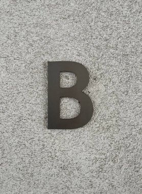 Açık gri desenli bir duvara monte edilmiş siyah B harfi güçlü bir kontrast ile minimalist ve modern bir kompozisyon oluşturur.