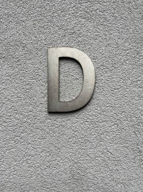 Metalik D harfi açık gri desenli bir duvara monte edilerek, temiz çizgiler ve ince kontrast ile minimalist ve modern bir kompozisyon oluşturuldu.
