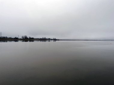 Görüntü, sakin ve pürüzsüz bir suyla sakin bir göl ya da nehir manzarasını tasvir ediyor. Uzak kıyı şeridinde, kırsal ya da yarı kırsal bir alanı işaret eden ağaçlar ve elektrik hatları bulunuyor. Kalın bir bulut katmanıyla bulutlu bir gökyüzü sessiz ve sakin bir atmosfer yaratır.
