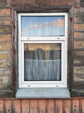 Beyaz çerçeveli, dantel perdeli bir pencereye yakın plan, eskimiş ahşap bir duvara yerleştirilmiş, rengi solmuş, mavi gökyüzünü ve bulutları yansıtan.