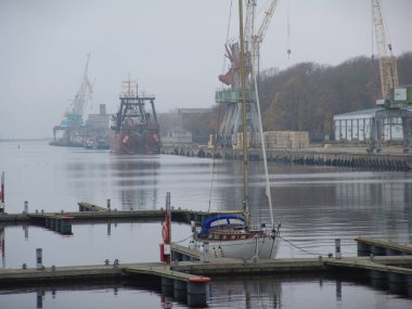 Ön planda demirli yelkenli, vinçler ve sakin nehir boyunca kargo gemileri olan sisli bir sanayi limanının yakın görüntüsü..