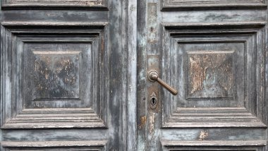 Yıpranmış ahşap çift kapılı, süslü panelleri olan, gri ve mavi tonlarında boyaları soyan, anahtar deliği olan eski bir metal saplı, yaş ve patina izleri gösteren bir kapı..