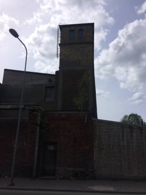 Uzun tuğla kulesi, metal merdiveni ve sokak lambası olan eski sanayi binası bulutlu bir gökyüzüne kurulmuş, kentsel bozulmayı ve tarihi mimariyi yakalıyor..