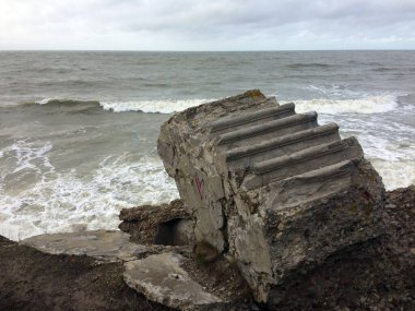 Bulutlu kıyı bozunması: Merdiven benzeri deseni ve grafitisi olan büyük, devrilmiş beton bir yapı, dalgaların bulutlu bir gökyüzü altında üzerine çarpmasıyla çökmekte olan bir set üzerine oturur..