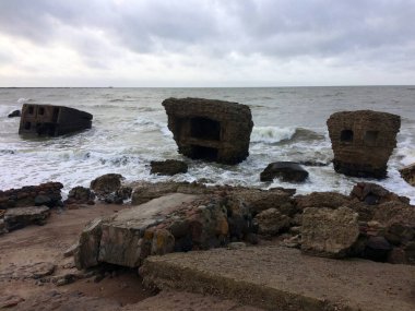 Bulutlu kıyı kalıntıları: Büyük beton sığınaklar dramatik, bulutlu bir gökyüzü altında kumlu ve kayalık bir sahilde dalgalar tarafından hırpalanmış..