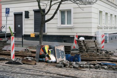 Demiryolu bakım sahnesi tramvay rayları üzerinde metal parçaları kaynak yapan bir işçiyle. Koruyucu teçhizat, araçlar, bariyerler ve dağılmış kaldırım taşları kentsel bir endüstriyel atmosfer yaratıyor..