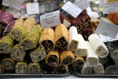 Bir market tezgahındaki tepside duran bir çeşit nougat dürümü. Çeşitli çeşitleri arasında beyaz nugat, fındık karışımları ve meyve dolu seçenekler yer alıyor. Görünür etiketler lezzet bilgisi sağlar.
