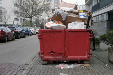 Kaldırımda duran inşaat atıkları, mukavva ve köpük malzemeleriyle dolu büyük kırmızı atık konteynırı. Park halindeki arabalar caddede, apartmanlar da arka planda. Bulutlu aydınlatma dağınık görünümü vurgular.