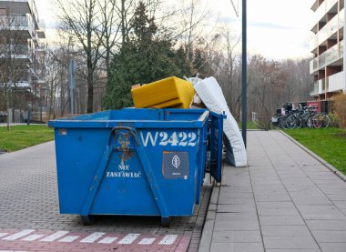 Kaldırıma yerleştirilmiş eski mobilyalar ve hantal eşyalar taşıyan mavi bir endüstriyel atık konteynırı. Doğal ışık, metal, kumaş ve asfalt dokularını ortaya çıkarır. Kompozisyon günlük şehir imha sahnesini belgeliyor..