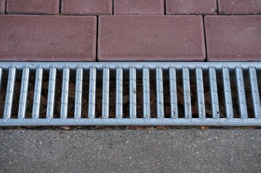 Kaldırımdaki asfalt yüzeyler arasına metal bir drenaj ızgarası yerleştirilmiş. Görüntü doğrusal desenlere, dokulara ve sessiz tonlara odaklanır. Doğal gün ışığı, metal ve taş malzemeler arasındaki zıtlığı vurgular..