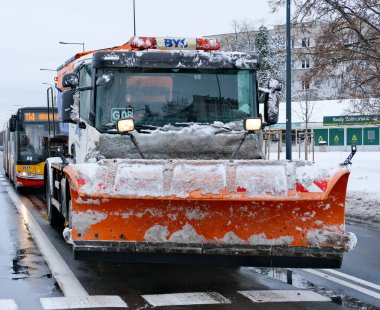 Bir kar temizleme aracının şehir yolundaki karları temizleme görüntüsü. Kar ve buz saban bıçağı ve araç yüzeyini kaplarken, toplu taşımacılık arkasında görünüyor. Doğal kış ışığı ve belgesel kompozisyonu şehir bakımını vurguluyor