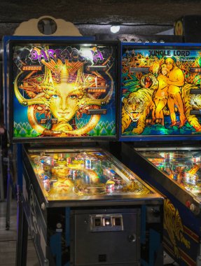 Barracora ve Orman Lordu 'nun arka camları olan iki eski tilt makinesi bir oyun salonunda yan yana duruyor. Aydınlatılan oyun alanları yansıtıcı cam altında tampon, rampa ve hedefler gösteriyor. İçerideki sıcak ışık doymuş fanteziyi güçlendirir..