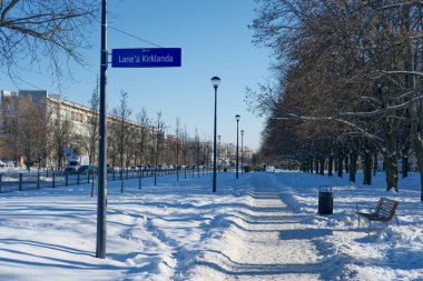 Şehir parkında üzerinde Skwer Lanea Kirklanda yazan mavi bir sokak tabelası olan karlı yaya geçidi. Uzun sokak lambaları, çıplak ağaçlar ve apartman blokları ve arabaları olan çitli bir yol arasındaki temiz bir yol. Parlak kış güneşi ışığı.