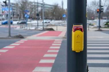 Sarı yaya geçidi düğmesi şehrin kavşağındaki karanlık bir direğe monte edilmiş. Aygıtta görünen CZEKAJ (WAIT) sözcüğüyle kırmızı ışıklı sinyal, yaya geçidi işaretleri ve arka planda bulanık trafik ışıkları.