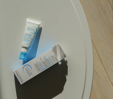 Nemlendirici jel krem tüpü ve güçlü güneş ışığı altında yuvarlak beyaz bir masaya yerleştirilmiş ürün kutusu. Kozmetik ambalajlar pürüzsüz yüzeyde keskin gölgeler bırakıyor. Yumuşak nötr tonlara sahip minimal iç ayarlar