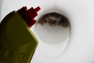 Klozet kapağına kırmızı açılı bir hortumla sarı bir temizleme şişesinin yakın çekimi kahverengi mineral birikimini gösteriyor, alçalmayı ve leke çıkarmayı gösteriyor..