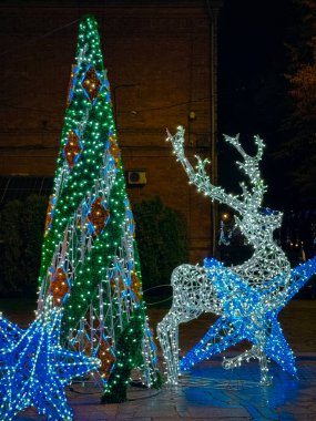 Açık havada LED rengeyiği, uzun ışıklı koni şeklindeki koni ağacı ve geceleri tuğla bir duvara karşı parlayan mavi yıldızların yer aldığı bir Noel süslemesi..