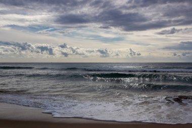Sonsuz deniz bulutlu bir gökyüzünün altında, dalgalı deniz dalgaları ve kumlu sahilde köpüklü sörf, dramatik deniz manzarası.