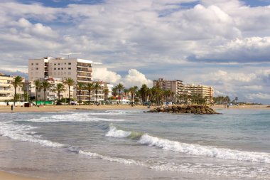 İspanya, Cunit 'daki kumlu sahilin panoramik manzarası, yumuşak deniz dalgaları, palmiye çizgili gezinti güvertesi ve dramatik bulutlu bir gökyüzü altında konut binaları.