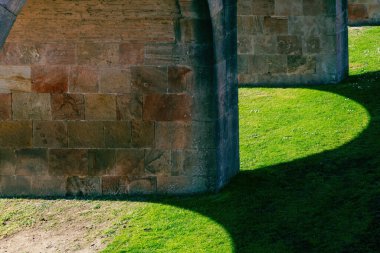 Yıpranmış taş kemerler ve tuğlalar güneş ışığında parlak yeşil çimenler üzerinde kıvrımlı gölgeler oluşturuyordu..