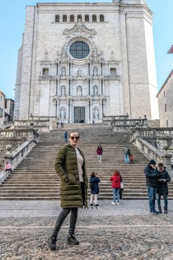 İkonik 91 taş basamak Girona Katedrali 'ne çıkıyor. Süslü Barok dış görünüşüyle. Yeşil kışlık ceketli kadın kaldırım taşı meydanında poz veriyor ve turistler devasa merdivenleri keşfediyor. Girona, İspanya - 28 Kasım 2021