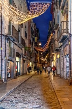 Akşam saatlerinde Girona Eski Kasabası 'ndaki Carrer de les Ballesteries caddesinin dikey çekimi. Noel ışıkları dar bir ortaçağ sokağında asılı, insanlar yürüyor ve alışveriş yapıyor. Girona, İspanya - 27 Kasım 2021