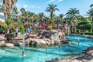 Alanya 'daki Kedi Evi Parkı' nın turkuaz fıskiyeleri ve gür palmiye ağaçları manzarası. Turistler aydınlık bir günde ünlü belediye kedi parkında yürürler. Alanya, Türkiye - 25 Ekim 2020