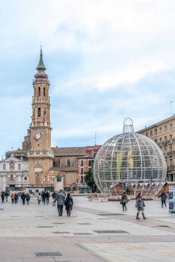 Yayalar tarihi La Seo Katedrali 'nin yanındaki meydanı geçip dev bir Noel süsü takıyorlar. Turistler Plaza del Pilar 'daki kış atmosferinin tadını çıkarıyorlar. Zaragoza, İspanya - 29 Kasım 2021