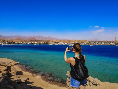 Genç kadın turist, Sharm el-Maya Körfezi 'nin Sharm El Sheikh' teki (Mısır) manzarasının fotoğraflarını çekiyor. Sırt çantası ve telefonu olan yetişkin bir kız Kızıl Deniz 'in arka planında bir kayanın üzerinde duruyor.