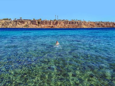 Bir kız Mısır 'ın Sharm El Sheikh kentindeki bir çöl kayasının arka planında Kızıl Deniz' in masmavi sularında yüzüyor. Genç bir kadın, egzotik bir tropik manzaranın arka planında rahatlıyor.