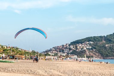 Alanya 'daki ünlü Kleopatra Sahili' nde iki kişilik paraşüt aktivitesi. Kumlu sahilde gökyüzünde mavi paraşüt insanlarla, güneşliklerle ve şehir binalarıyla. Türkiye - 25 Ekim 2020