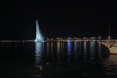 Cenevre 'deki Jet d' Eau çeşmesinin göz kamaştırıcı bir gece manzarası. Karanlık sulara yansıyan parlak şehir ışıkları ve ön planda sakin ve atmosferik bir şehir manzarası yaratan bir tekne..