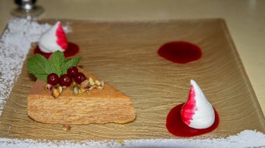 Fıstık, fındık ezmesi ve nane şekeriyle süslenmiş katmanlı kek ve altın desenli bir tabakta sanatsal olarak hazırlanmış çilek sosu. Zarif bir tatlı, güzel yemekler ve pastane konsepti.