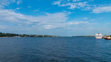 Uzak kıyı şeridi, küçük kasaba evleri ve dramatik bulutların altında rıhtıma yakın beyaz bir yolcu gemisi ile geniş bir yaz manzarası. Seyahat ve doğa geçmişi.
