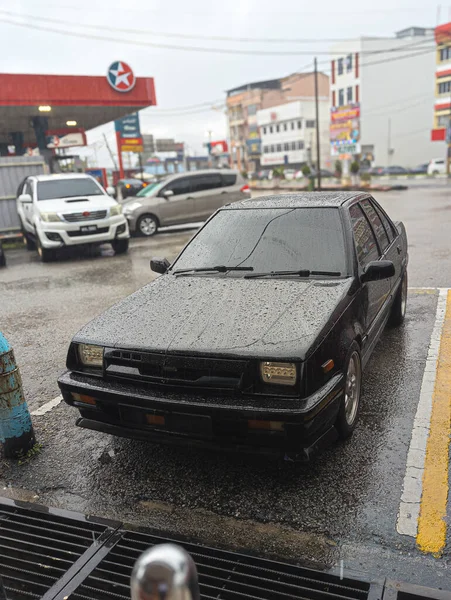 Malezya. 27 Kasım 2025. Bir Texaco istasyonunda yağmurlu bir gün ıslak bir şehir parkına park etmiş eski model siyah bir sedan nostaljik titreşimler uyandırıyor..