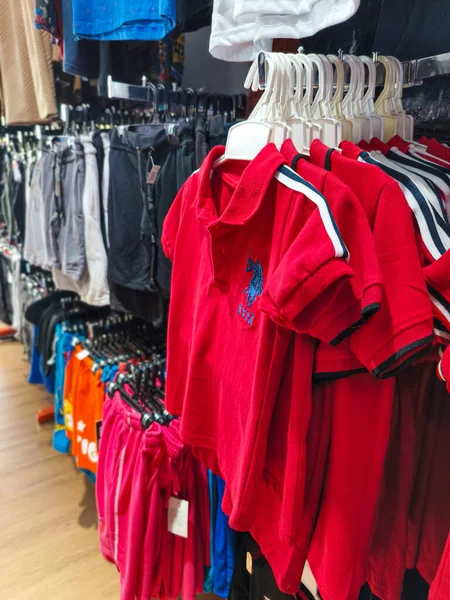 Malezya. 13 Mart 2025. Mavi logolu bir sıra kırmızı polo gömlek, diğer renkli kıyafetlerle birlikte bir mağazanın rafında asılı..