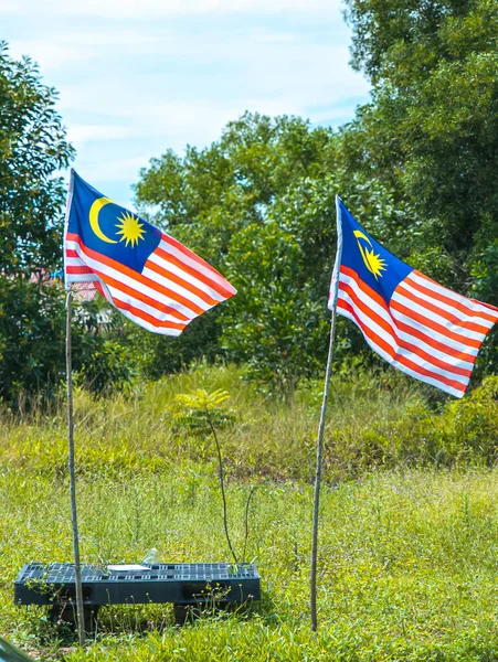 İki Malezya bayrağı (Jalur Gemilang) açık mavi gökyüzünün altında yemyeşil ağaçlarla dolu çimenli bir alanda ince kutuplarda dalgalanıyor..