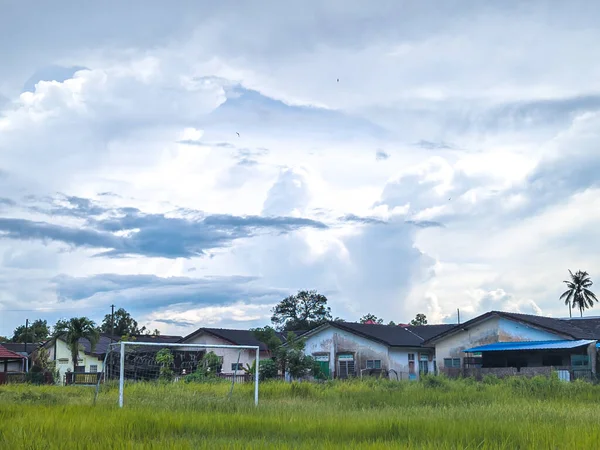 Futbol kalesine sahip çimenlik bir alan dramatik bulutlu gökyüzünün altında mütevazı binaların önünde duruyor..