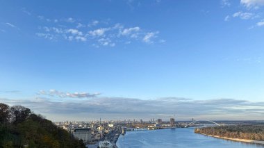 Açık mavi gökyüzü altında nehir kıyısı, köprü ve sakin su yansıması olan geniş panoramik bir şehir. Kopyalama alanı olan en küçük pankart arkaplanı.