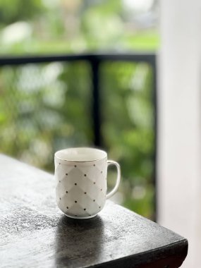 Küçük siyah noktalardan oluşan elmas ızgaralı beyaz seramik kupa, bulanık yeşil arka plan ve siyah metal parmaklıklar ile karanlık bir yüzeye yerleştirilmiş, sakin bir açık veranda düzeneğini işaret ediyor..