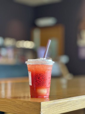 Johor, Malezya, 10 Kasım 2025: Sade bir plastik fincanda kırmızı buzlu meyve çayı, üzerinde kahve çekirdeği ve aromalı arpa logosu bulunan ve ahşap bir kafe masasına yerleştirilmiş..