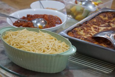 Fırınlanmış lazanya, makarna, et sosu, rendelenmiş peynir, meyve salatası ve cam bir masada satay..